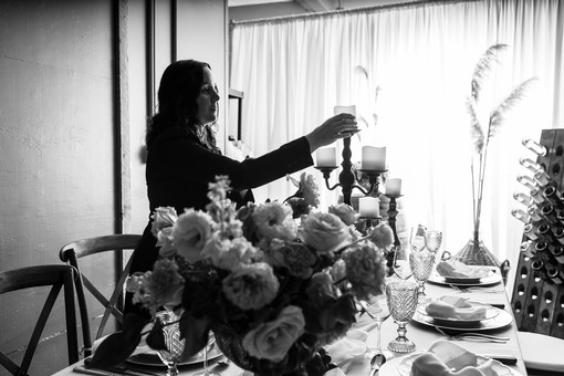 black and white image of woman lighting candles on a table with a vase of flowers.