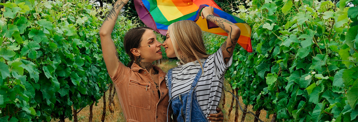 Image of two women embracing in a row of wine vines while holding up a rainbow flag